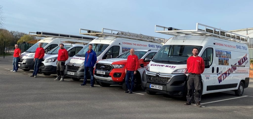 Five Window-Mart uPVC installersin matching red shirts stand beside four branded vans with ladders in a Horsham car park.