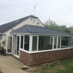 A single-storey West Sussex house with a modern brick and glass conservatory extension featuring a dark tiled roof, uPVC white window frames, and a glass door, set beside a paved path and surrounded by grass and trees.
