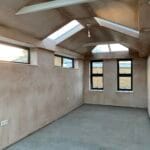 Unfurnished room in West Sussex with freshly plastered walls, large uPVC windows, two rooflights, and a concrete floor. Sunlight streams in, highlighting the modern, spacious, and unfinished interior.
