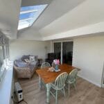 A bright sunroom with large uPVC windows and a skylight, featuring a wooden dining table with four chairs, a cosy seating area with sofas.