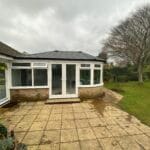 A small brick and glass conservatory with uPVC double doors opens onto a stone patio, surrounded by a grassy garden with a large tree and shrubs in West Sussex on a cloudy day.
