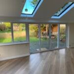 A bright room with wooden floors, large uPVC floor-to-ceiling glass doors, and two skylights. The doors open to a patio and a green garden with trees, a fence, and a wooden shed in the background—perfect for West Sussex living.