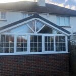 A brick and glass conservatory with uPVC white-framed windows is attached to the back of a two-storey house with a tiled roof in West Sussex, under a partly cloudy sky.