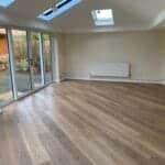 A bright, empty room with light brown wood flooring, cream walls, and large uPVC glass doors opening to a garden. Skylights and recessed lights illuminate the space. Located in West Sussex, it features a white radiator and glass-panelled door.