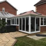 A brick house in West Sussex with a modern glass conservatory extension featuring a red tiled roof, uPVC white-framed windows, and sliding doors, opening onto a paved patio with some greenery and an outdoor hot tub.