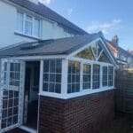 A brick and glass conservatory extension with white uPVC-framed windows and a grey tiled roof is attached to a house in West Sussex. The glass doors are open, and the extension sits beside a fenced garden area.