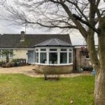A single-storey West Sussex house with a uPVC conservatory featuring large windows and a dark roof, set in a back garden with a lawn, patio, potted plants, and a large tree in the foreground.