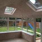 A bright conservatory with uPVC windows, tiled floors, cream-coloured walls, and skylights in the ceiling. Outside, in West Sussex, there’s a green lawn, a blue slide, a shed, and a wooden fence.