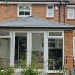 A modern brick house in West Sussex with a newly built white uPVC conservatory featuring large glass doors and windows, a dark grey roof, and a fenced back garden with plants and a small patio area.