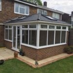 A modern uPVC brick and glass conservatory with a dark tiled roof is attached to the back of a house in West Sussex, featuring large windows and a patio area with garden ornaments and potted plants.