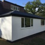 A modern uPVC house extension with white exterior walls, black trim, large windows, and a dark roof, attached to a brick home in West Sussex, surrounded by trees under a partly cloudy sky.