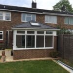 A brick house in West Sussex featuring a white-framed uPVC glass conservatory extension in the back garden, adjacent to a wooden fence and a small patio area with potted plants on a grassy lawn.