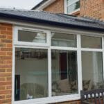 A brick conservatory with large uPVC white-framed windows and a black roof is attached to a house in West Sussex. Inside, there is a wicker chair and sofa with cushions.