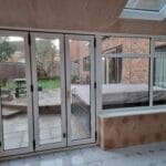 View from inside a conservatory with tiled floor and uPVC glass doors looking out onto a garden with outdoor seating and a covered hot tub next to a brick house in West Sussex.