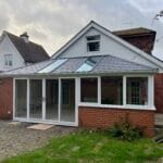 A brick house in West Sussex with a white-framed uPVC glass conservatory extension, featuring three roof windows and large sliding glass doors, opening onto a paved patio bordered by grass and plants.