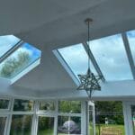 A modern conservatory with large uPVC glass roof panels, white walls, and a geometric star-shaped pendant light. Outside, there is a green garden with trees, a swing set, and a wooden bench in tranquil West Sussex.