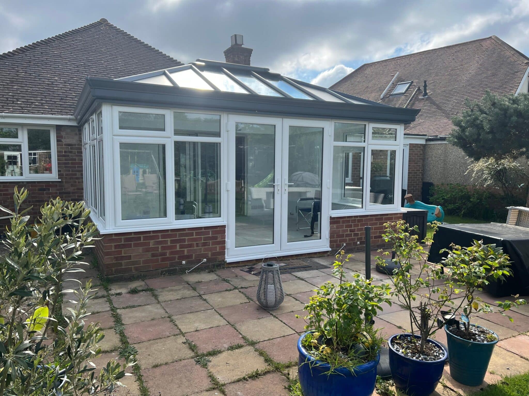 A modern glass conservatory with a brick base and sleek uPVC frames is attached to a house in Horsham. There are potted plants on a tiled patio in the foreground.