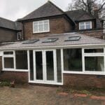 A brick house in West Sussex with a single-storey uPVC extension featuring three rooflights, large windows, and double glazed doors opening onto a paved patio. The sky is overcast and there are trees in the background.