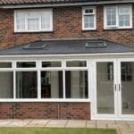 A modern brick house extension in West Sussex with large uPVC white-framed windows and a glass door, featuring a dark tiled roof with two rooflights, opening onto a paved patio and grassy lawn.