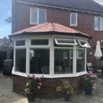 A brick house in West Sussex with a hexagonal uPVC conservatory featuring large windows and a red tiled roof. Potted pink hydrangeas decorate the stone patio, while the partly sunny garden enjoys a blue sky.
