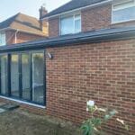 A newly built brick all-season room in West Sussex features large uPVC sliding glass doors, a flat tiled roof, and modern wall light.