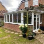 A brick house in West Sussex with a white-framed uPVC glass conservatory, surrounded by a garden with potted plants, a wooden pergola, and a paved path on a grassy lawn. Neighbouring houses are visible in the background.