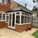 A small uPVC conservatory with white frames and a dark roof is attached to a red-brick house in West Sussex. It features large windows and glass doors opening onto a patio with potted flowers and artificial grass nearby.