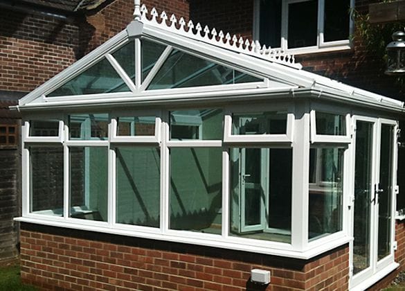 Bifold and Patio Door Installation A white uPVC-framed glass conservatory with a brick base is attached to a red brick house in Horsham. The conservatory features large windows, a triangular glass roof section, and decorative trim along the roof edge.