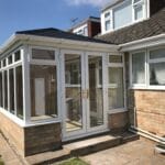 A modern brick and glass conservatory extends from a house in West Sussex, featuring white-framed uPVC windows and double glazed doors opening onto a stone patio with a small garden area nearby under a clear blue sky.