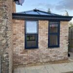 Small brick extension with a dark-framed glass roof, two blue uPVC-framed windows, and a paved area in front, attached to an older brick building with a satellite dish in West Sussex.