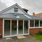A modern house extension in West Sussex with large uPVC glass sliding doors, brick walls, and a sloping roof featuring rooflights. The extension opens onto a small paved patio area with grass and plants nearby.