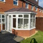 A small brick and glass conservatory with a tiled roof is attached to the back of a West Sussex house. It features uPVC white window frames, a wooden decking, a grassy area, and a wooden fence nearby.