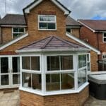 A brick house in West Sussex with a uPVC white-framed conservatory featuring large windows and a brown tiled roof, set on a paved patio with a hot tub and outdoor furniture visible.