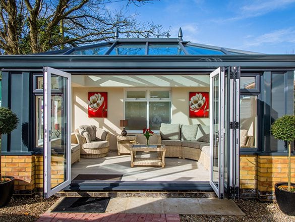 A modern uPVC conservatory in Billingshurst with a glass roof, open double doors, rattan furniture, a beige sofa, red flower wall art, and two potted topiary trees on either side of the entrance.