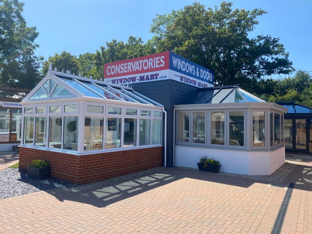 Two uPVC glass conservatories on display outside a building with a CONSERVATORIES WINDOWS & DOORS WINDOW-MART sign in Horsham. The sunny area features trees in the background and potted plants by the entrance.