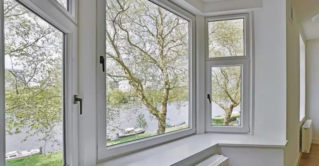 Cost Of UPVC Windows West Sussex With Installation, Large uPVC bay windows with white frames overlook a river and leafy trees on a bright day in Horsham. A white windowsill and part of a radiator are visible inside.