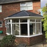 A brick house in West Sussex with a glass conservatory extension featuring a dark tiled roof, uPVC white window frames, and a small garden area in the foreground.