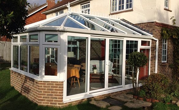 A white uPVC-framed glass conservatory extends from a brick house in West Sussex, featuring sliding doors, windows, and a pitched glass roof. Inside are chairs, a table, and plants, with grass and shrubs surrounding the space.