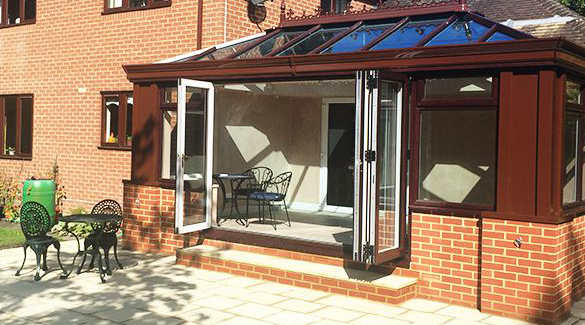 A brick conservatory with uPVC-framed glass roof and large open French doors connects to a house in Horsham. Black metal garden furniture sits outside on a paved area, with a green water butt visible nearby.
