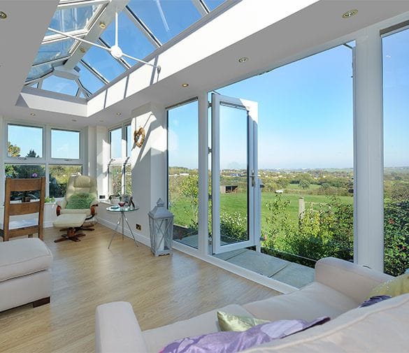 A bright sunroom in Horsham with large uPVC glass windows and a glass ceiling, featuring light sofas, a wooden chair, and green countryside views under a clear blue sky.