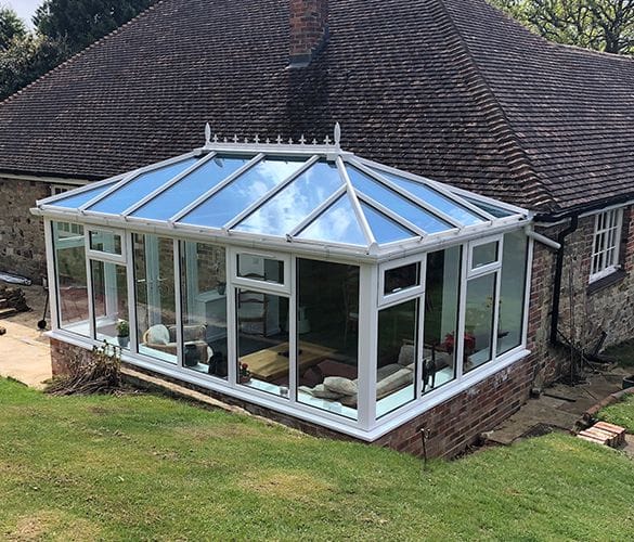 A modern uPVC and glass conservatory with a white frame is attached to a brick house in Horsham, featuring a sloped glass roof and large windows. Inside, furniture is visible. The conservatory overlooks a grassy garden.