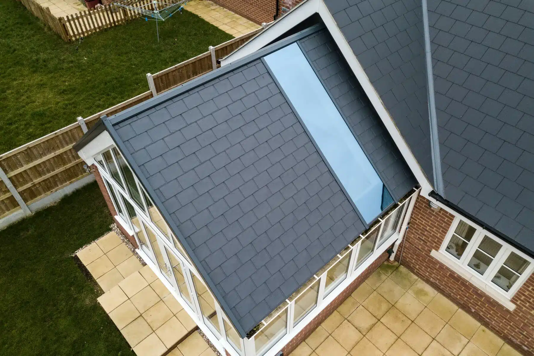 Aerial view of a house extension in Horsham with a grey tiled roof, large glass skylight, and uPVC features, adjacent to a brick building with a fenced garden and paved patio area.