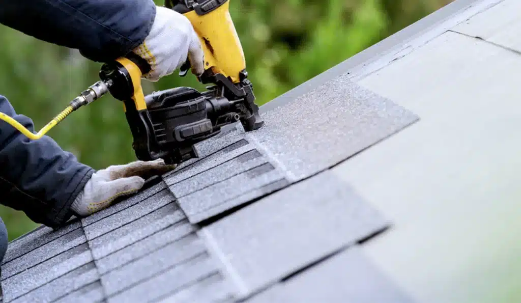 Conservatory Roof Replacement Horsham, with A Window Mart installer in gloves uses a nail gun to secure asphalt shingles onto a sloping roof, preparing or repairing the roofing with precise placement on a Horsham property.