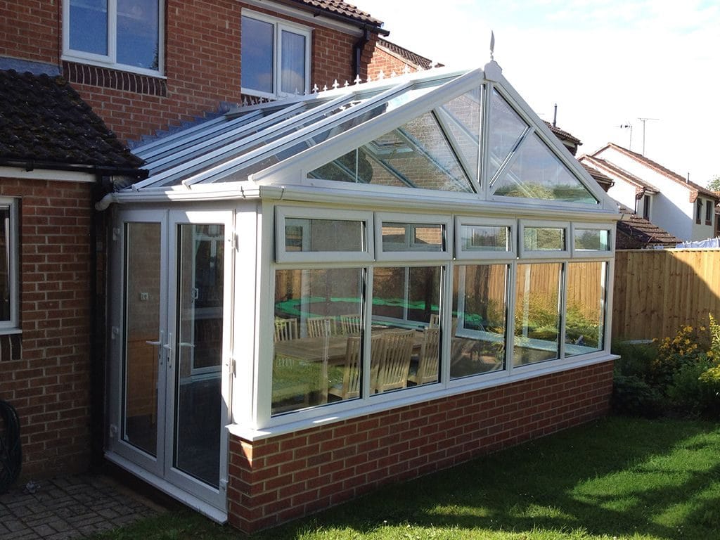 A glass conservatory with a pitched roof and white uPVC frame is attached to a house. Featuring large windows and a glass door overlooking a green lawn and wooden fence.