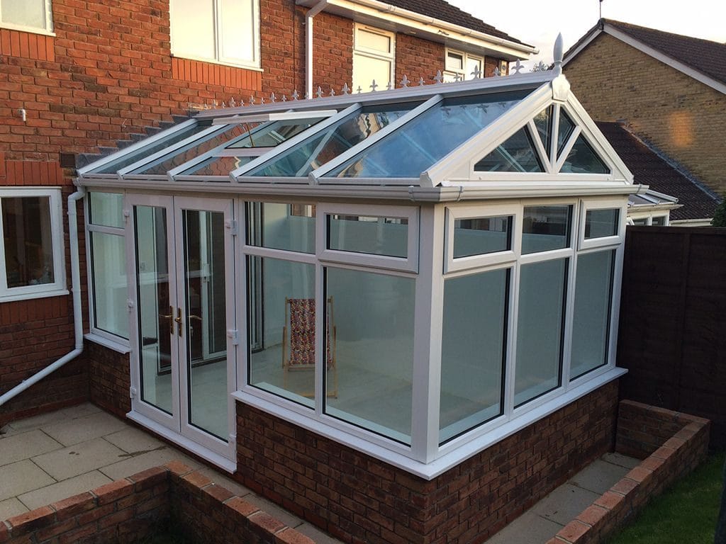 A modern uPVC glass conservatory with a white frame and pitched roof is attached to a house. The lower panels are frosted glass, and the tiled floor leads to double doors opening onto a small paved patio.