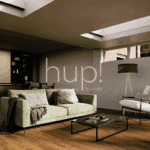 A modern Horsham Hup! extension with a green sofa, armchair, standard lamp, and coffee table on wooden flooring; skylights in the ceiling and kitchen cupboards are visible in the background.