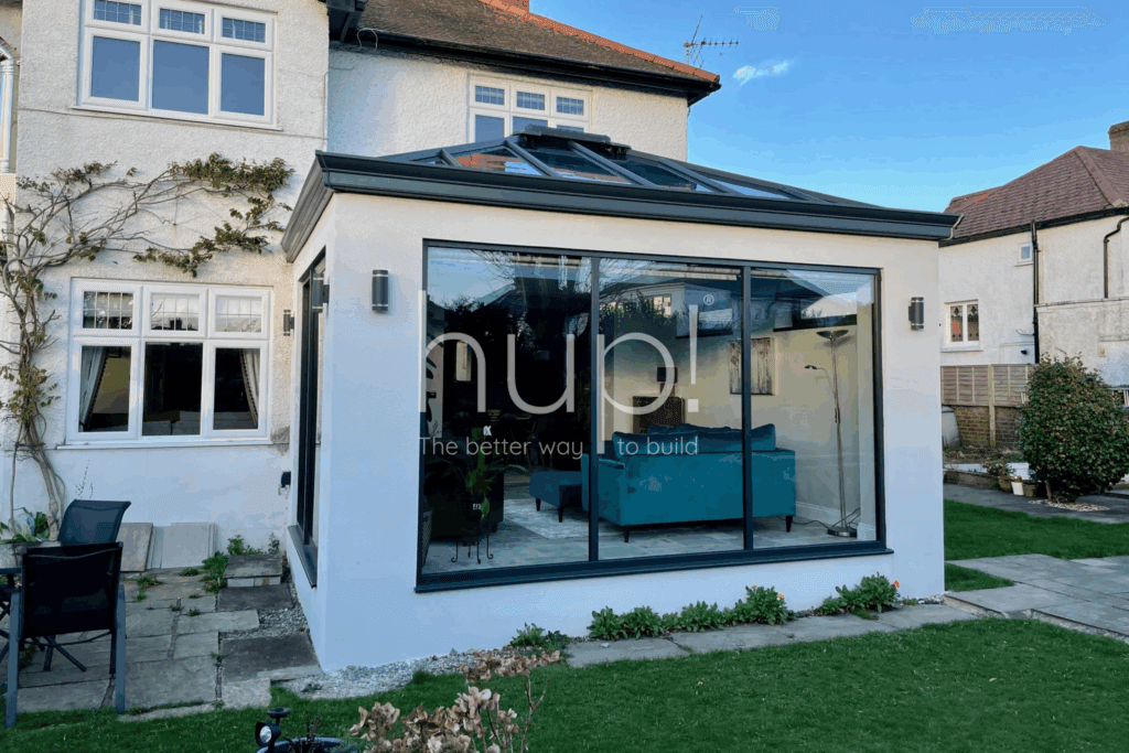 A modern Hup! extension with a blue settee and green chairs attached to a white house in Horsham, surrounded by a garden and patio furniture.