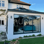 A modern Hup! extension with a blue settee and green chairs attached to a white house in Horsham, surrounded by a garden and patio furniture.