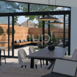 Modern dining area with a dark table and chairs, a laptop, and pendant lights, opening to a patio with outdoor seating and greenery through large uPVC glass doors.