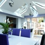 A modern dining area with a white table, purple and black chairs, indoor plants, and skylights. Large uPVC windows and glass doors let in sunlight, revealing an outdoor patio with a table and parasols in Horsham.